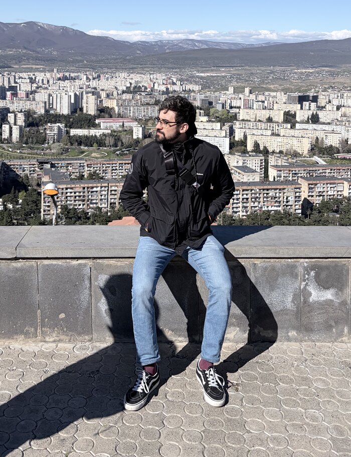 Thiago Lavado sentado num muro alto, de jaqueta preta, com a cidade de Tbilisi e as montanhas ao fundo.